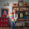 Ein Mädchen mit geflochtenen Zöpfen hält eine bunte Einhorn-Schultüte und lächelt fröhlich vor einer Kulisse mit großen Buchstaben, Büchern und Schulmotiven – perfekte Erinnerung zur Einschulung in Karlsruhe.