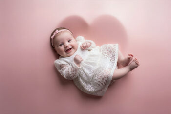 Lachendes Baby im weißen Spitzenkleid beim Babyfotoshooting auf rosa Hintergrund mit Herzform im Fotostudio von Belly Deluxe in Karlsruhe
