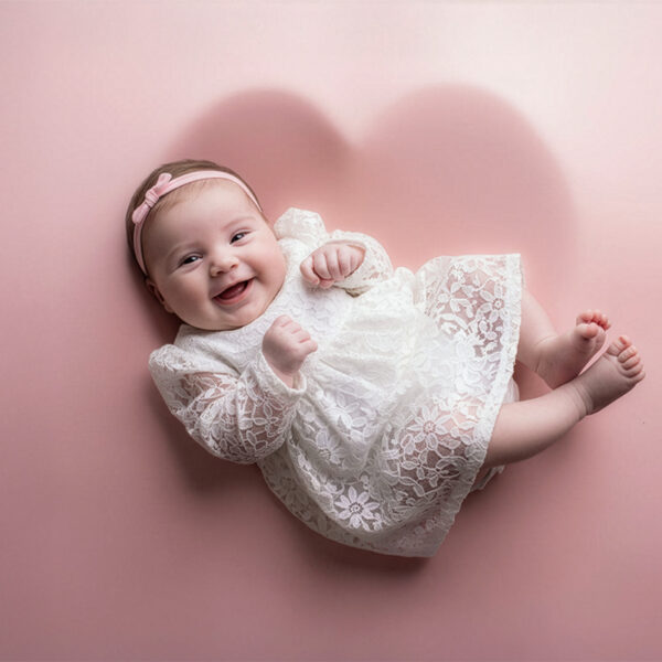Lachendes Baby im weißen Spitzenkleid beim Babyfotoshooting auf rosa Hintergrund mit Herzform im Fotostudio von Belly Deluxe in Karlsruhe