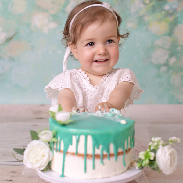 Cake-Smash-Fotoshooting in Karlsruhe – Baby im weißen Kleid sitzt hinter einer Torte mit türkisfarbener Glasur und Blumen und lächelt.