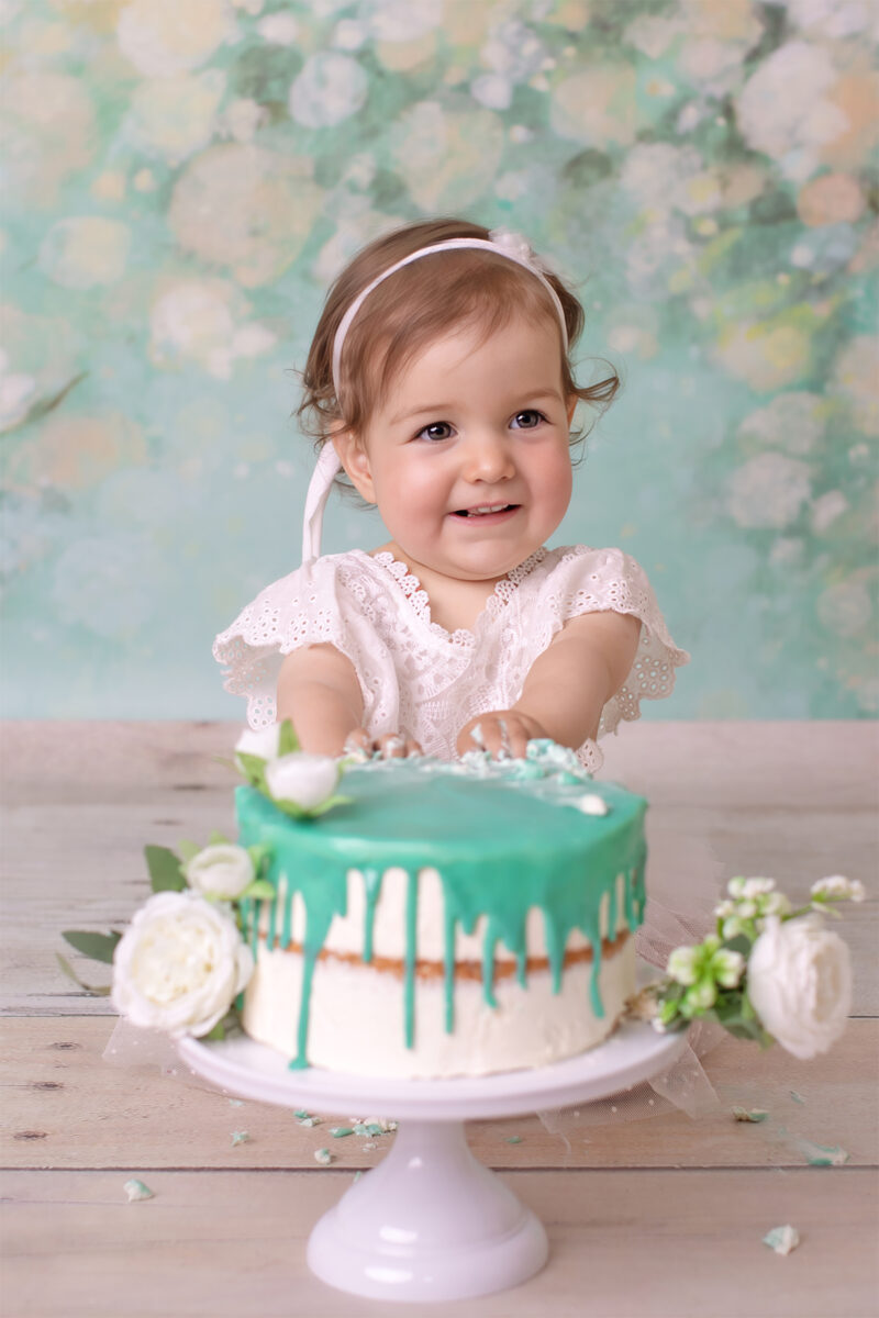 Cake-Smash-Fotoshooting in Karlsruhe – Baby im weißen Kleid sitzt hinter einer Torte mit türkisfarbener Glasur und Blumen und lächelt.
