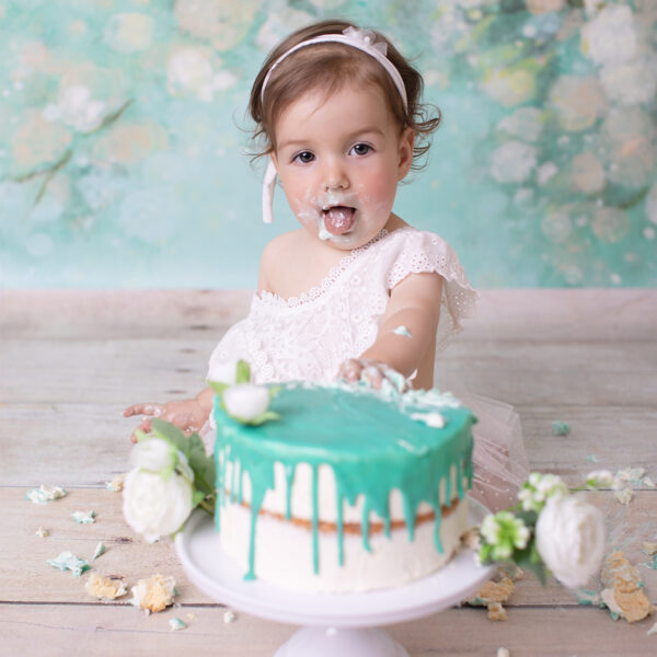 Cake-Smash-Fotoshooting in Karlsruhe – Baby mit Creme am Mund sitzt hinter einer Torte mit türkisfarbener Glasur und steckt die Zunge raus.