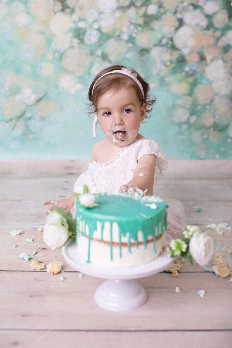 Cake-Smash-Fotoshooting in Karlsruhe – Baby mit Creme am Mund sitzt hinter einer Torte mit türkisfarbener Glasur und steckt die Zunge raus.
