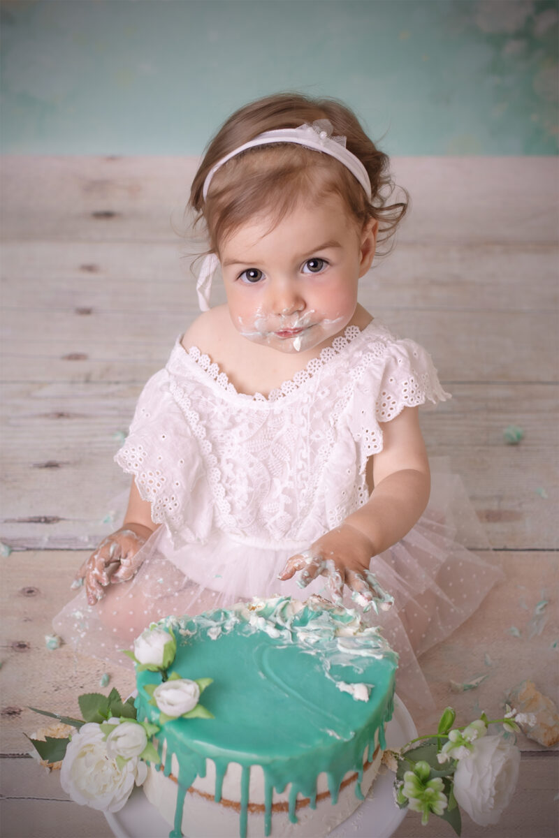 Cake-Smash-Fotoshooting in Karlsruhe – Baby im weißen Kleid sitzt vor einer mintfarbenen Torte mit Blumen und hat Creme im Gesicht.