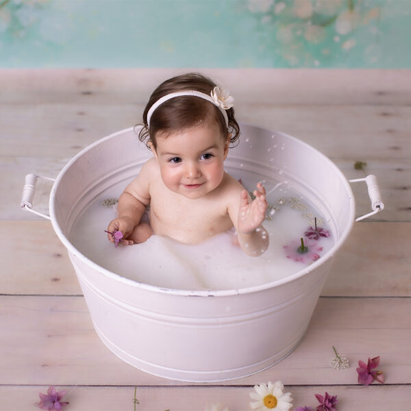 Kind sitzt im Milchbad in einer weißen Wanne im Fotostudio, zarte Blüten schwimmen im Wasser.