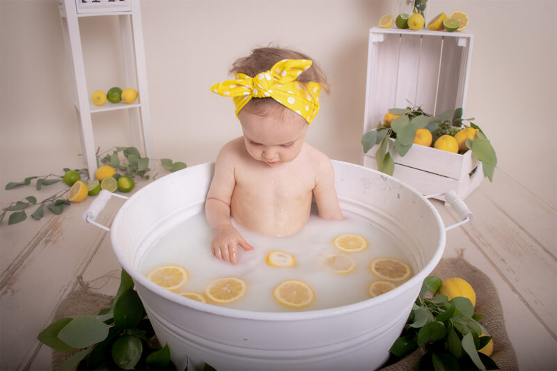 Kind sitzt im Milchbad in einer weißen Wanne und spielt mit Zitronenscheiben im Wasser, im Set liegen Zitronen und Limetten.
