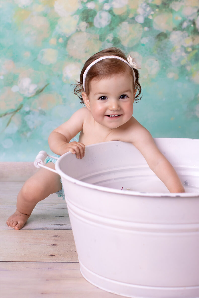 Kind lehnt an einer weißen Wanne im Fotostudio und lächelt in die Kamera, pastellfarbener Hintergrund.