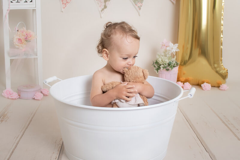 Kind sitzt in einer weißen Wanne im Fotostudio und kuschelt mit einem Teddy, im Hintergrund Deko und goldener Luftballon.
