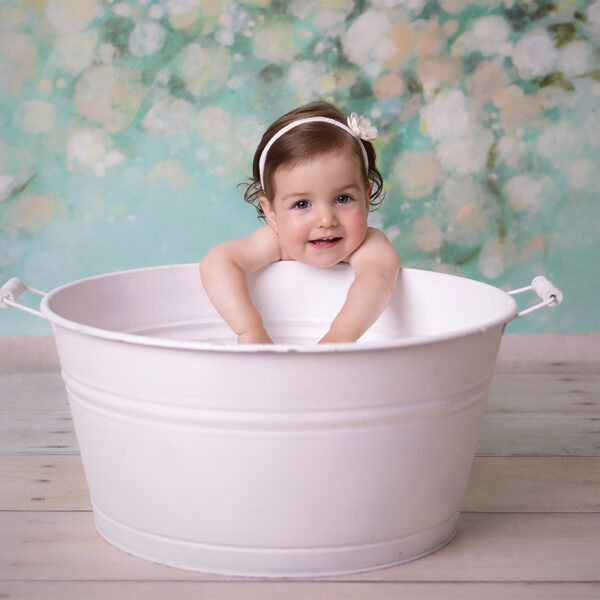 Kind lehnt in einer weißen Wanne im Fotostudio und lächelt in die Kamera, weicher Hintergrund in Pastellfarben.