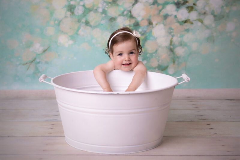 Kind lehnt in einer weißen Wanne im Fotostudio und lächelt in die Kamera, weicher Hintergrund in Pastellfarben.