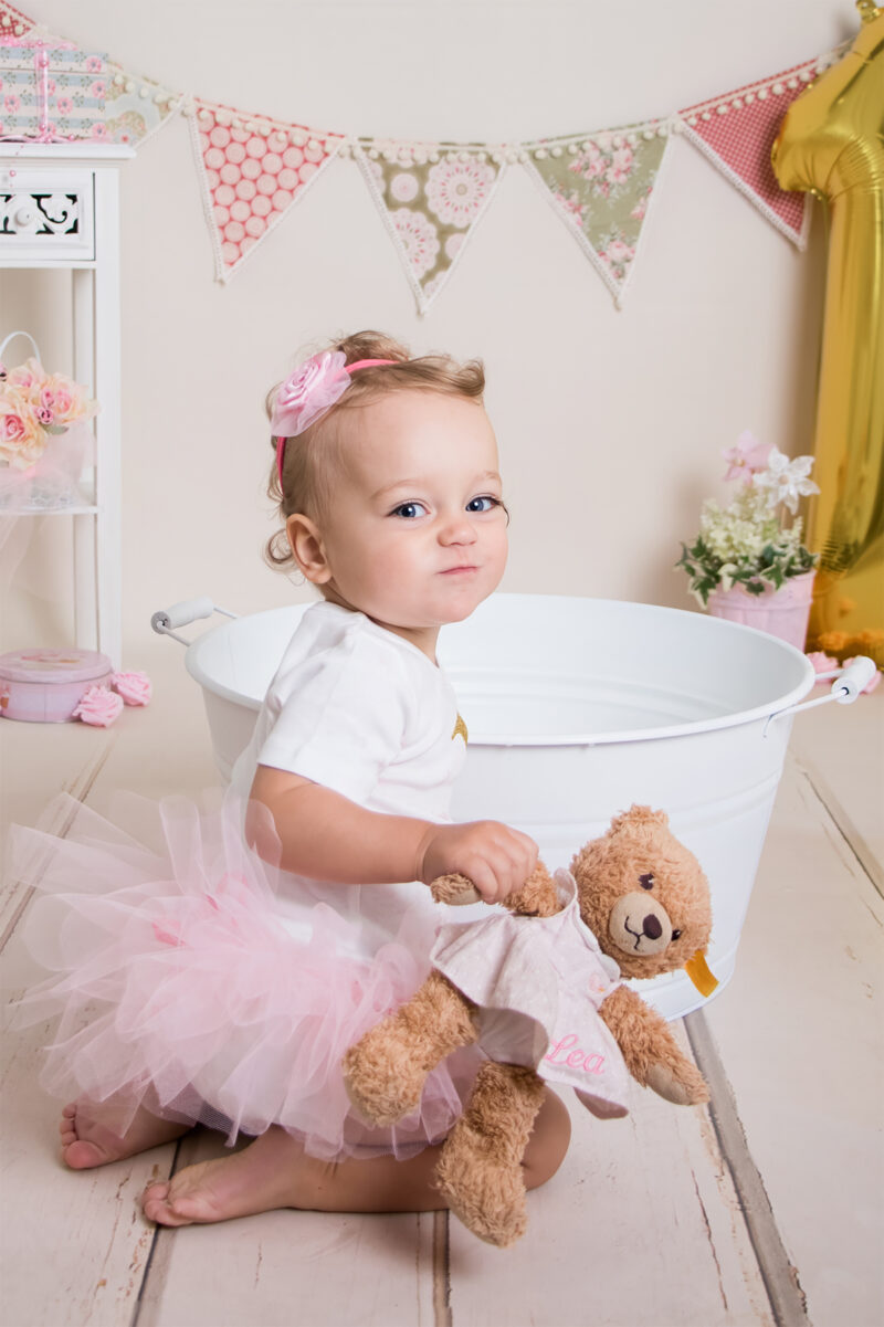 Kind sitzt im Fotostudio vor einer weißen Wanne und hält einen Teddy, im Hintergrund eine Wimpelkette und ein goldener Luftballon.