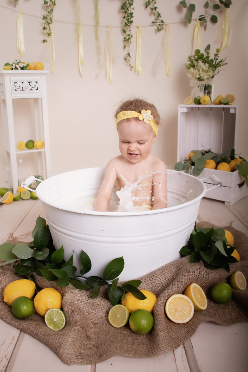 Kind sitzt im Milchbad in einer weißen Wanne, umgeben von Zitronen und Limetten im hellen Studio-Set.