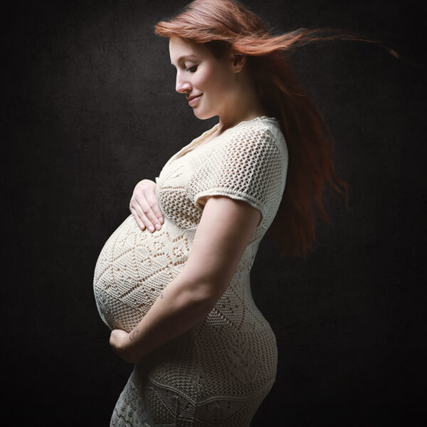 Kunstvolle Schwangerschaftsfotografie im Studio Karlsruhe vor dunklem Hintergrund, Profilpose mit sanftem Licht und Windmaschine, die Haare wehen nach hinten und setzen Bewegung.