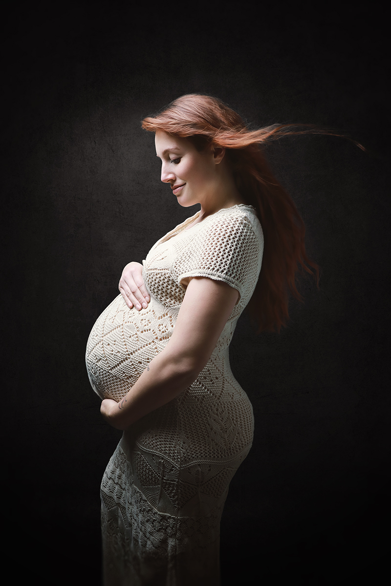 Kunstvolle Schwangerschaftsfotografie im Studio Karlsruhe vor dunklem Hintergrund, Profilpose mit sanftem Licht und Windmaschine, die Haare wehen nach hinten und setzen Bewegung.