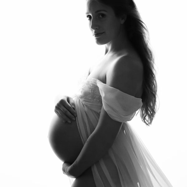Schlichte Schwarzweiß-Schwangerschaftsfotografie vor weißem Hintergrund, Schwangere im Profil mit fließendem Chiffonkleid, Hände am Babybauch.
