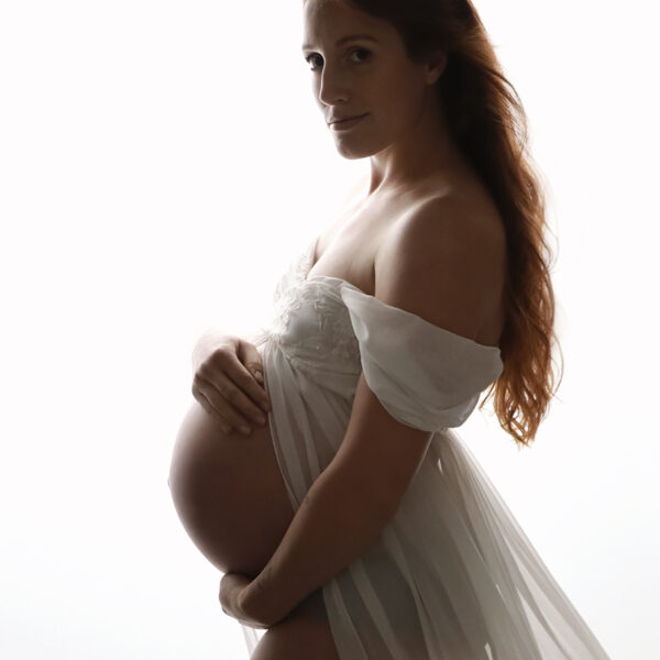 Fine-Art-Schwangerschaftsfotografie im Atelier Karlsruhe: Schwangere Frau im Profil vor weißem Hintergrund, trägt ein schulterfreies weißes Kleid mit transparentem, fließendem Stoff, hält den Babybauch mit beiden Händen und blickt ruhig in die Kamera; weiches Licht betont die zarte Silhouette.