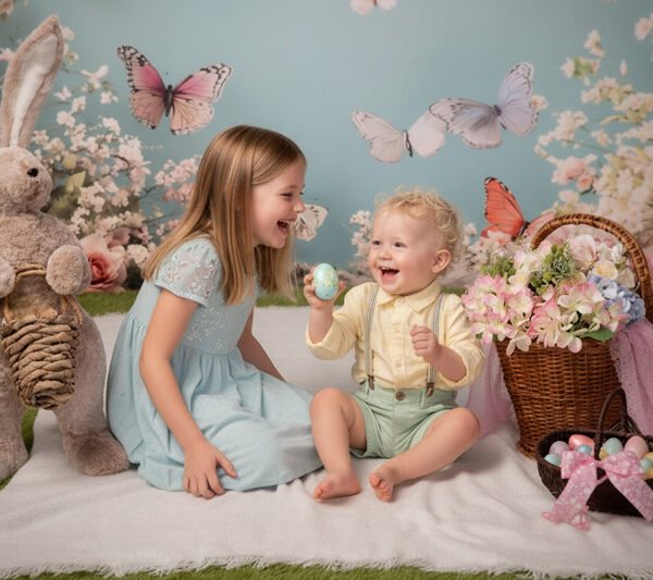 Zwei Kinder lachen im Oster-Set mit großem Hasen, Blumen, Körben und bunten Eiern vor pastelligem Hintergrund.