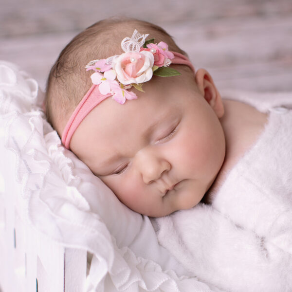 Schlafendes Baby mit rosa Blumen-Stirnband liegt eingekuschelt in weißer Decke in einem weißen Bettchen vor hellem Holzhintergrund.