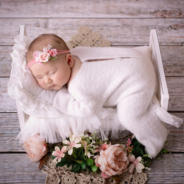 Schlafendes Baby mit rosa Blumen-Stirnband liegt in einem kleinen weißen Bettchen auf Holzplanken-Hintergrund, zugedeckt mit weißer Decke und umgeben von rosa Blumen.