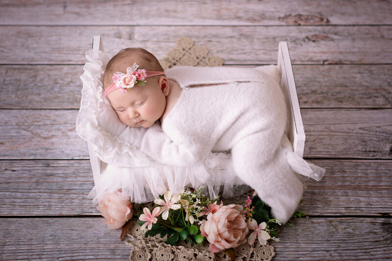 Schlafendes Baby mit rosa Blumen-Stirnband liegt in einem kleinen weißen Bettchen auf Holzplanken-Hintergrund, zugedeckt mit weißer Decke und umgeben von rosa Blumen.