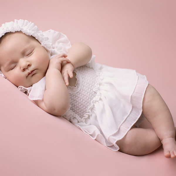 Schlafendes Baby im weißen Kleid und Häubchen liegt seitlich auf rosafarbenem Hintergrund, Hände unter dem Kinn, Beine leicht angewinkelt.