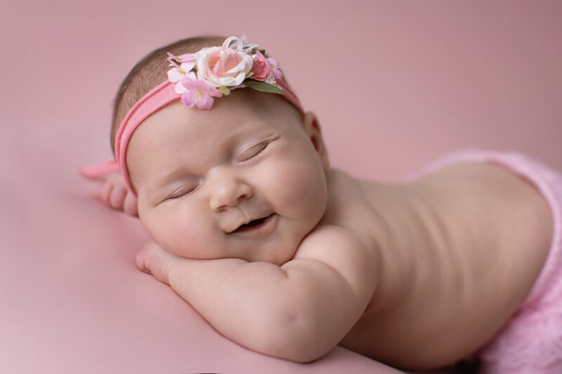 Lächelndes Baby liegt auf rosafarbenem Hintergrund, trägt ein rosa Stirnband mit Blumen und hat die Augen geschlossen.