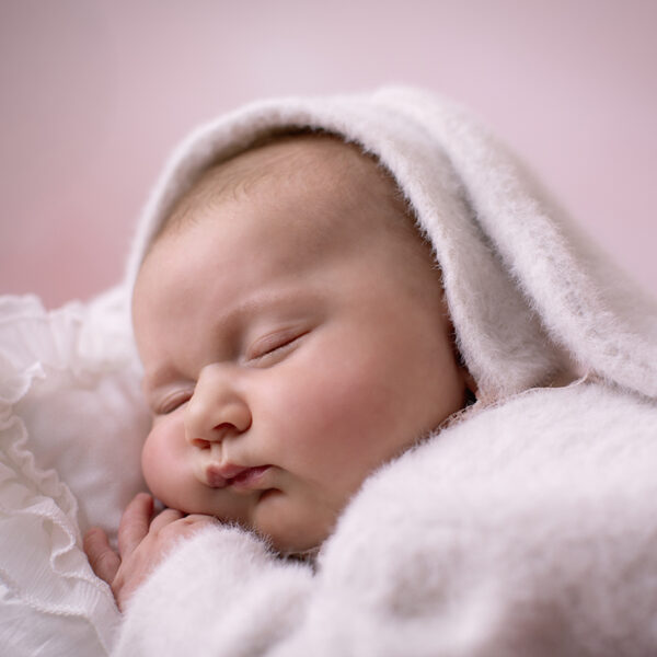 Schlafendes Baby im flauschigen Hasenkostüm mit Kapuze liegt seitlich in einem weißen Bettchen vor zart rosafarbenem Hintergrund.