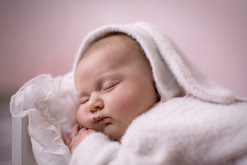 Schlafendes Baby im flauschigen Hasenkostüm mit Kapuze liegt seitlich in einem weißen Bettchen vor zart rosafarbenem Hintergrund.