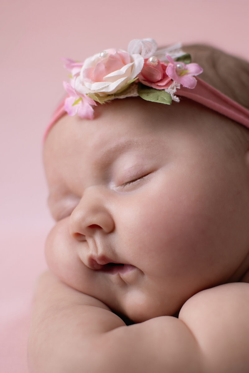 Nahaufnahme eines schlafenden Babys mit rosa Blumen-Stirnband vor rosafarbenem Hintergrund, Gesicht und Wange weich im Fokus.