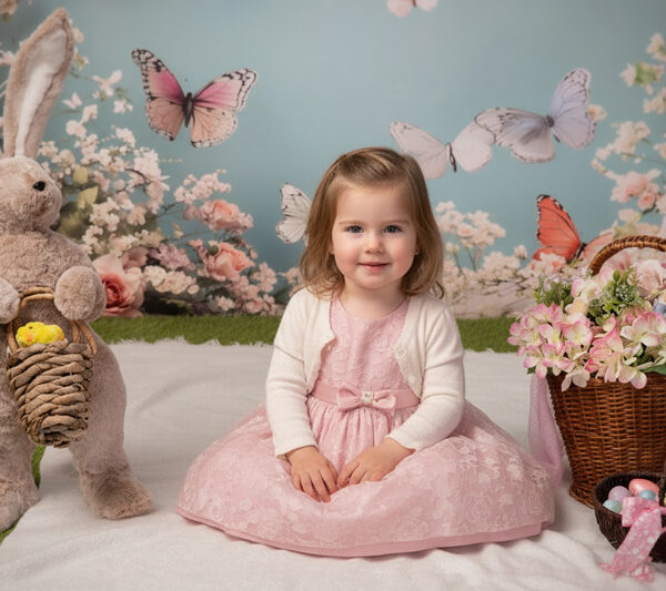 Kleines Mädchen sitzt im Oster-Set zwischen großem Plüschhasen, Blumenarrangements und Ostereiern vor pastelligem Hintergrund.