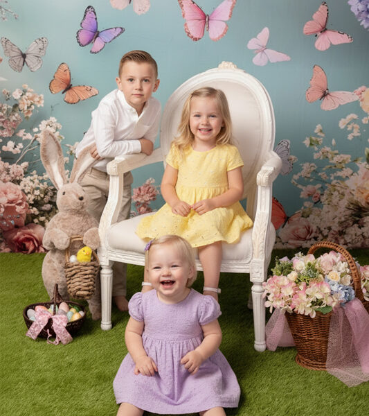 Drei Kinder im Oster-Set mit weißem Sessel, Schmetterlingskulisse, großem Hasen, Blumen und Ostereiern auf grünem Boden.