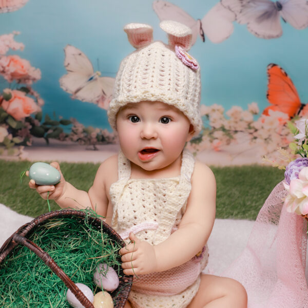 Baby im gehäkelten Osterhasen-Outfit sitzt im frühlingshaften Set mit Ostereiern und Körbchen und hält ein Ei in der Hand.
