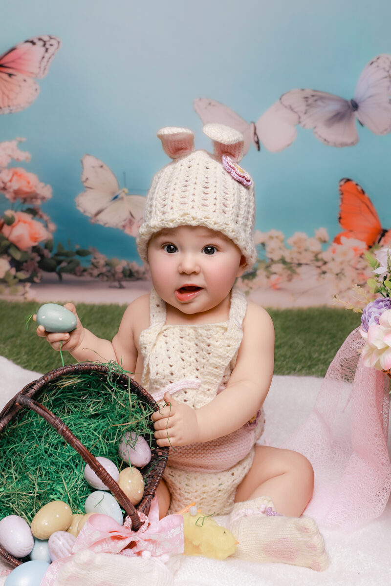 Baby im gehäkelten Osterhasen-Outfit sitzt im frühlingshaften Set mit Ostereiern und Körbchen und hält ein Ei in der Hand.