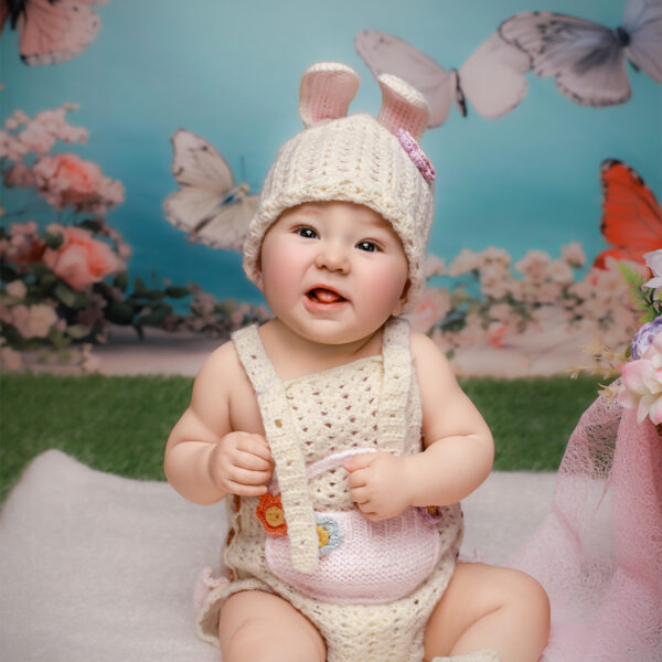 Baby im Osterhasen-Outfit sitzt auf einer Decke im frühlingshaften Set und lächelt in die Kamera, Hintergrund mit Schmetterlingen.