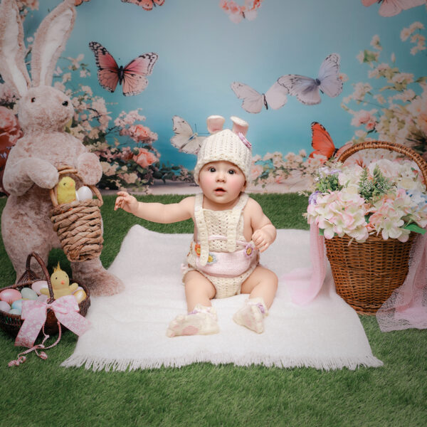 Baby im Osterhasen-Outfit sitzt im Oster-Set mit Hasenfigur, Ostereiern und Blumenkorb vor blauem Schmetterlingshintergrund.