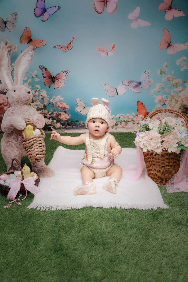 Baby im Osterhasen-Outfit sitzt im Oster-Set mit Hasenfigur, Ostereiern und Blumenkorb vor blauem Schmetterlingshintergrund.
