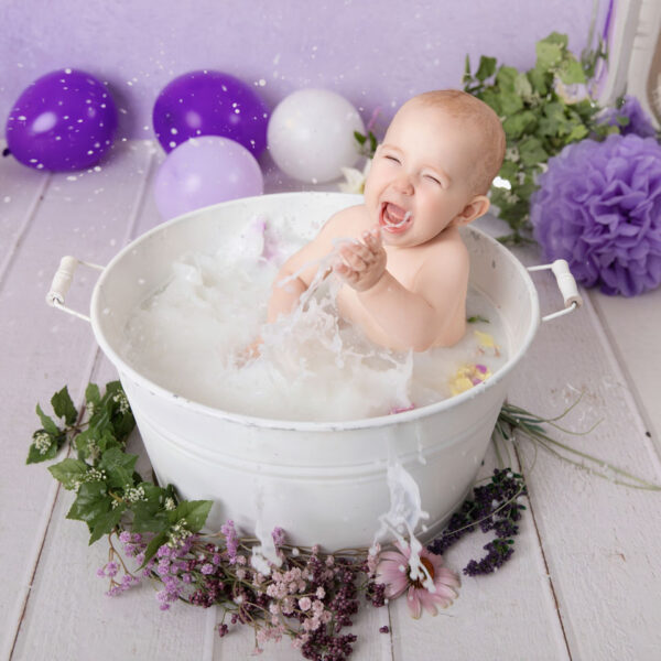 Baby lacht laut und spritzt im Milchbad in einer weißen Wanne vor lila Hintergrund, fotografiert im Studio von Belly Deluxe in Karlsruhe.