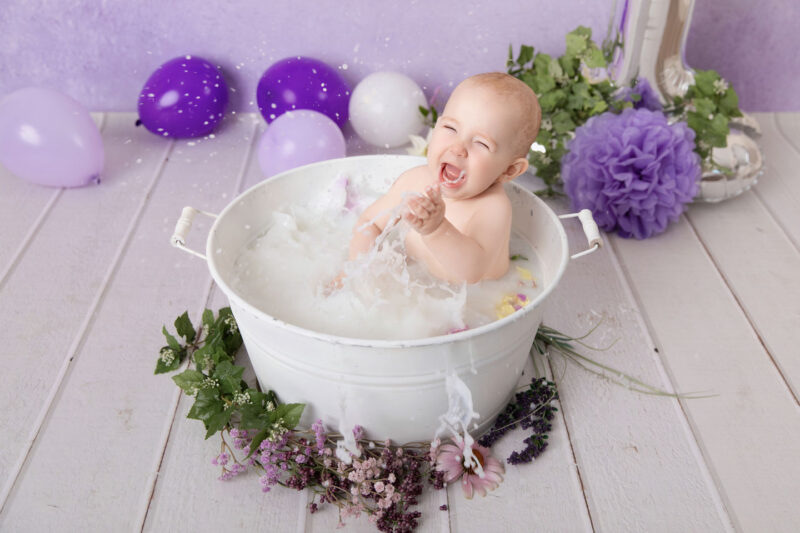 Baby lacht laut und spritzt im Milchbad in einer weißen Wanne vor lila Hintergrund, fotografiert im Studio von Belly Deluxe in Karlsruhe.