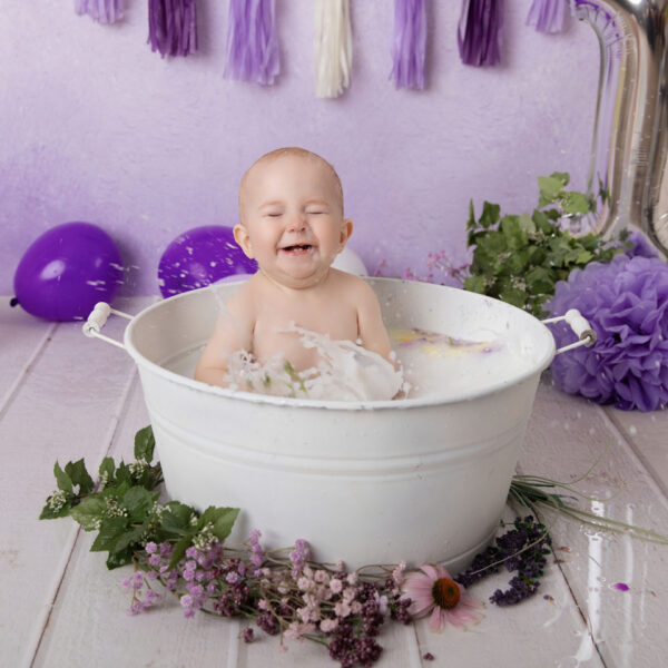 Baby lacht beim Planschen im Milchbad in einer weißen Wanne vor lila Hintergrund, fotografiert im Studio von Belly Deluxe in Karlsruhe.