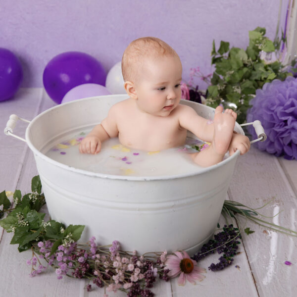 Baby sitzt im Milchbad in einer weißen Wanne vor lila Hintergrund und spielt mit dem Fuß, fotografiert im Studio von Belly Deluxe in Karlsruhe.