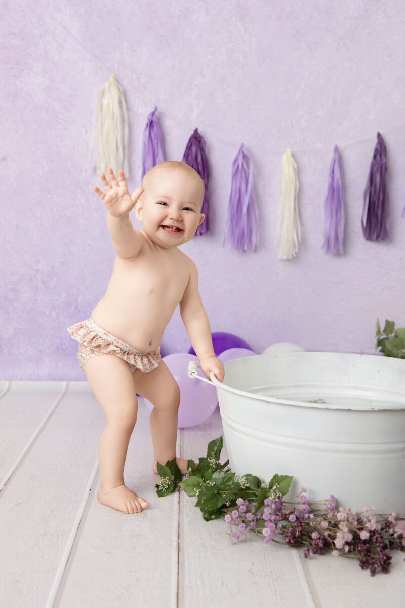 Baby steht neben der weißen Wanne und winkt vor lila Hintergrund mit Ballons, fotografiert im Studio von Belly Deluxe in Karlsruhe.