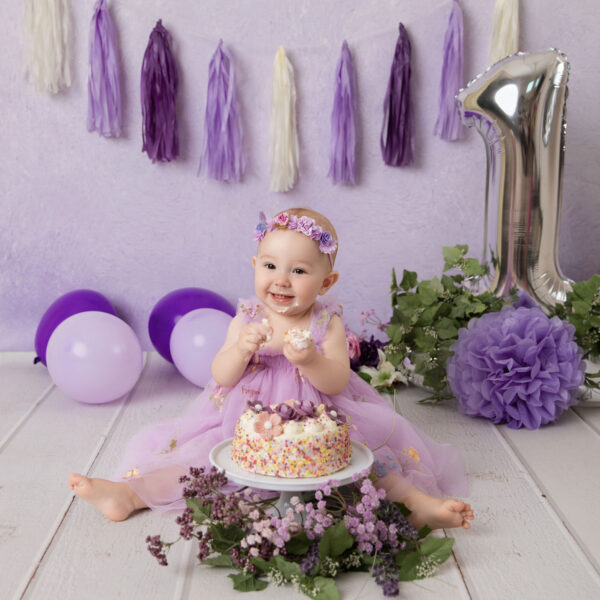 Baby im lila Kleid sitzt im Cakesmash-Set mit lila Hintergrund, Ballons, Quasten-Girlande und großer Zahl 1 vor einer Mini-Torte, fotografiert im Studio von Belly Deluxe in Karlsruhe.