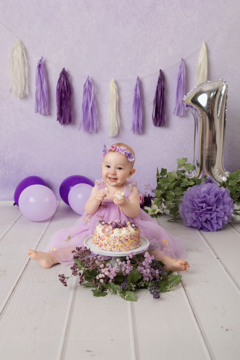 Baby im lila Kleid sitzt im Cakesmash-Set mit lila Hintergrund, Ballons, Quasten-Girlande und großer Zahl 1 vor einer Mini-Torte, fotografiert im Studio von Belly Deluxe in Karlsruhe.