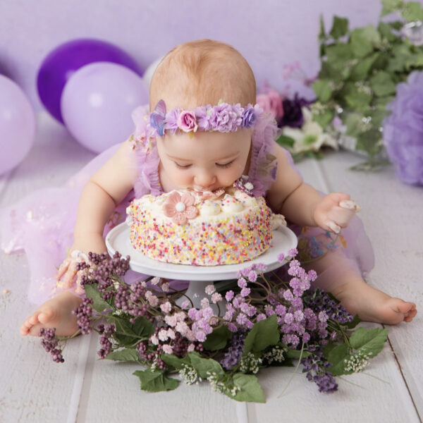 Baby im lila Kleid isst beim Cakesmash von einer bunten Mini-Torte vor lila Hintergrund, fotografiert im Studio von Belly Deluxe in Karlsruhe.