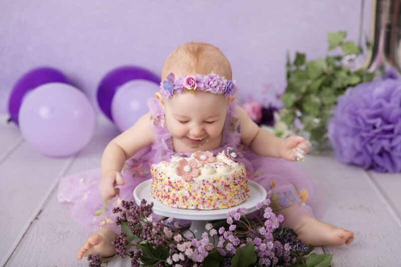 Baby im lila Kleid beugt sich lachend über eine bunte Mini-Torte beim Cakesmash vor lila Hintergrund, fotografiert im Studio von Belly Deluxe in Karlsruhe.