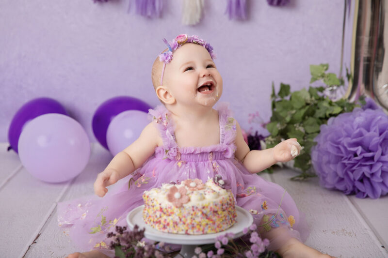 Baby im lila Kleid lacht beim Cakesmash vor lila Hintergrund mit bunter Mini-Torte, fotografiert im Studio von Belly Deluxe in Karlsruhe.