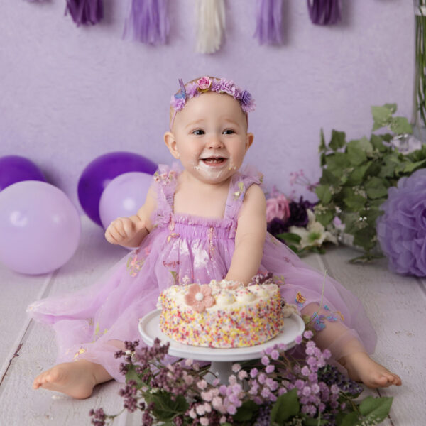 Baby im lila Kleid lacht beim Cakesmash vor lila Hintergrund mit Mini-Torte auf einem Kuchenständer, fotografiert im Studio von Belly Deluxe in Karlsruhe.