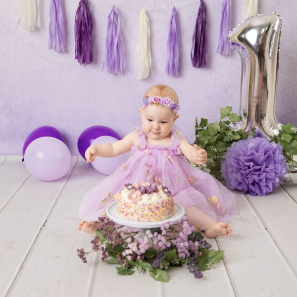 Baby im lila Kleid sitzt im Cakesmash-Set mit lila Hintergrund, Ballons und großer Zahl 1 neben einer Mini-Torte, fotografiert im Studio von Belly Deluxe in Karlsruhe.