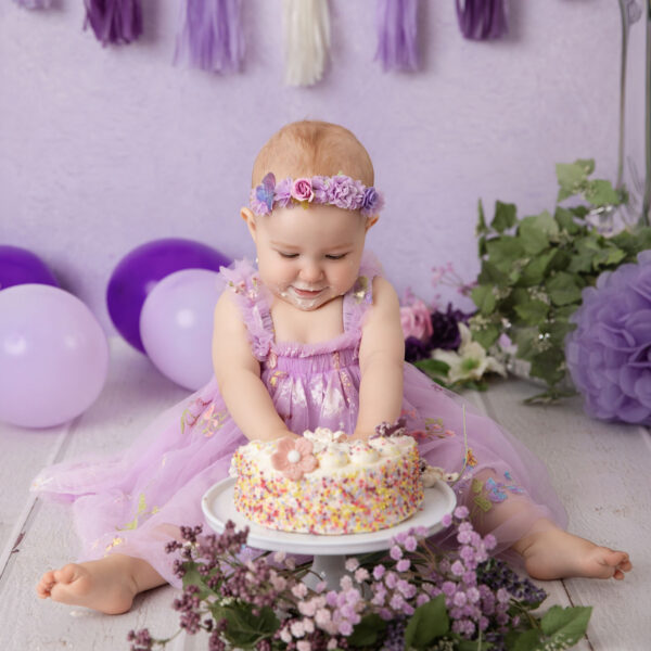 Baby im lila Kleid sitzt vor lila Hintergrund und greift nach einer bunten Mini-Torte beim Cakesmash, fotografiert im Studio von Belly Deluxe in Karlsruhe.