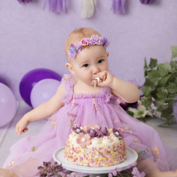 Baby im lila Kleid nascht beim Cakesmash von der Creme und sitzt vor lila Hintergrund mit Mini-Torte, fotografiert im Studio von Belly Deluxe in Karlsruhe.